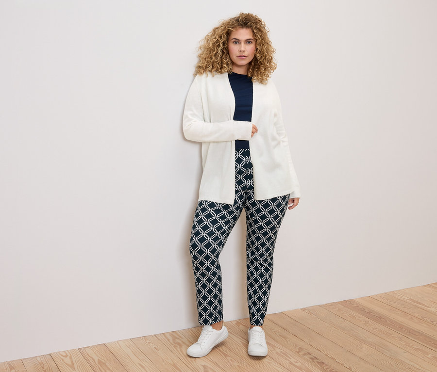 Žena s kudrnatými vlasy v bílém kardiganu, tmavě modré halence a vzorovaných kalhotách stojící před bílou stěnou.