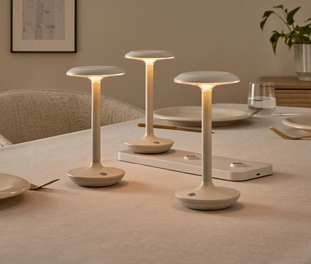 Tři minimalistické, krémové stolní lampy na prostřeném jídelním stole, jedna z nich na nabíjecí stanici. Lampy svítí teplým světlem.