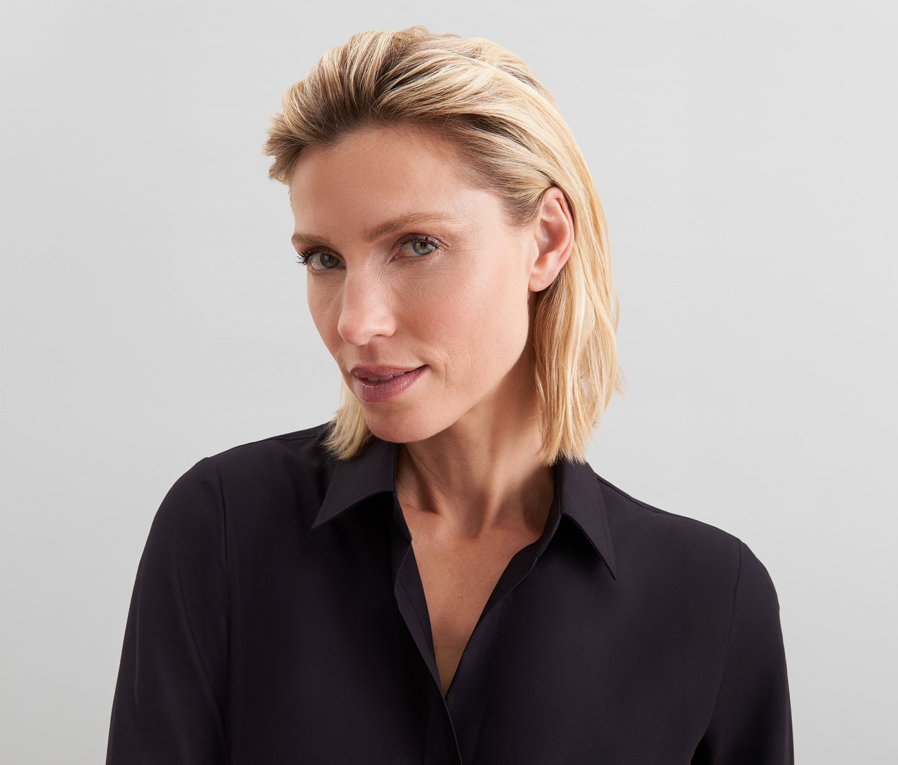 Detailní záběr ženy s krátkými blond vlasy, která má na sobě černou košili a dívá se mírně do strany.