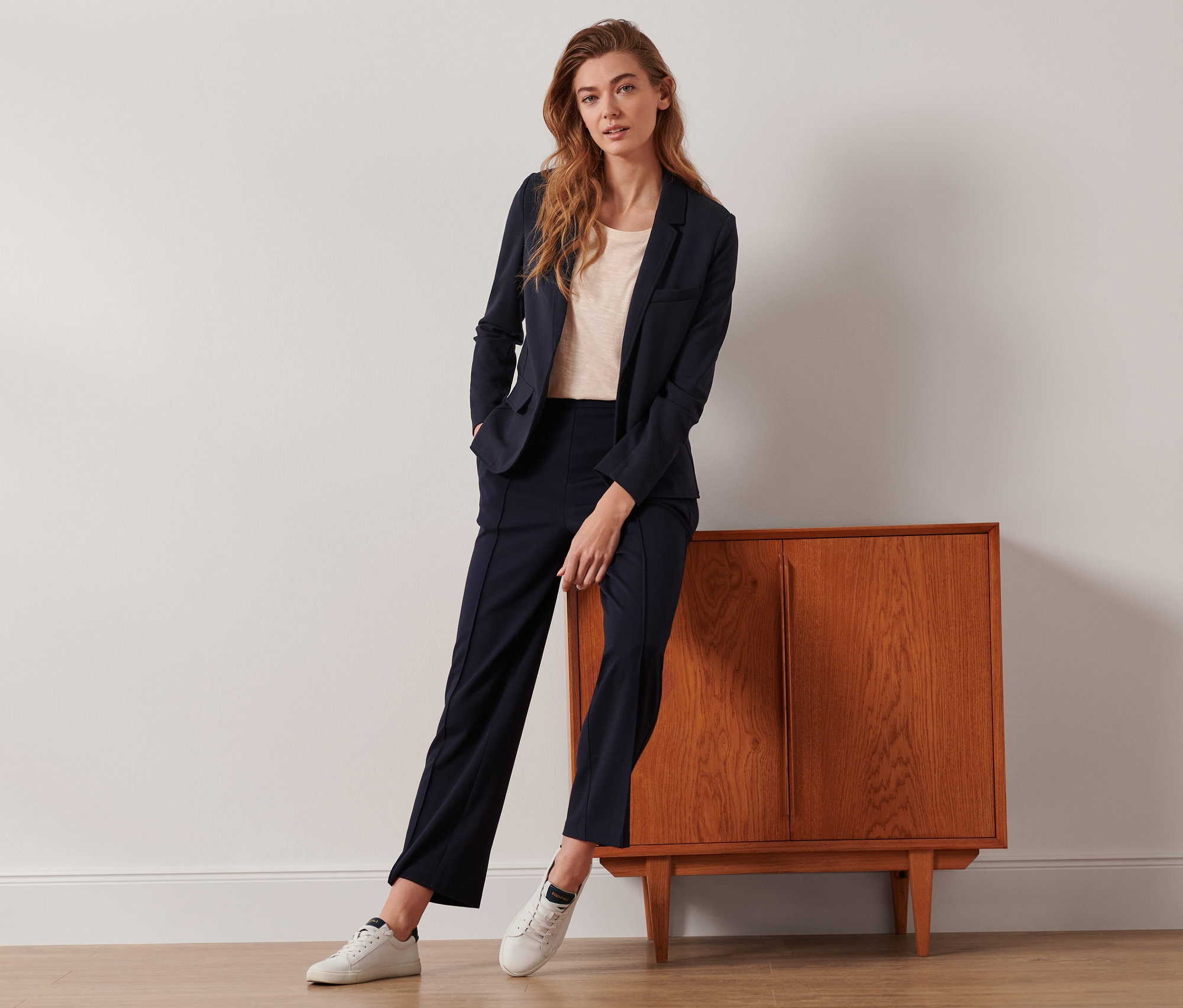 Žena pózuje opřená o skříňku v džerzejovém blejzru a tmavě modrých žerzejových kalhotách s puky.