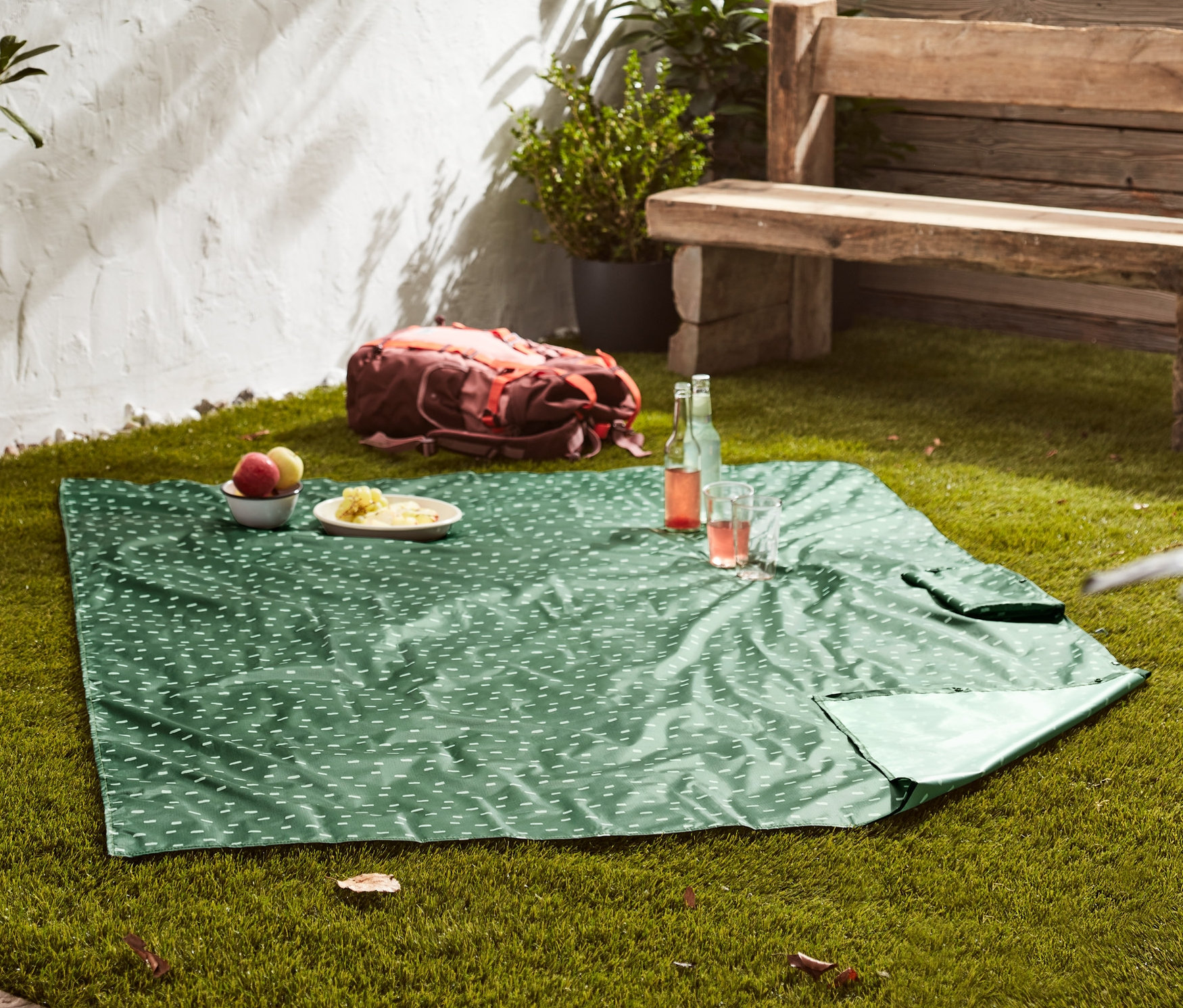 Zelená deka na piknik 2 v 1 s bílým vzorem leží na trávě. Na dece jsou položené talíře s jídlem a pitím.