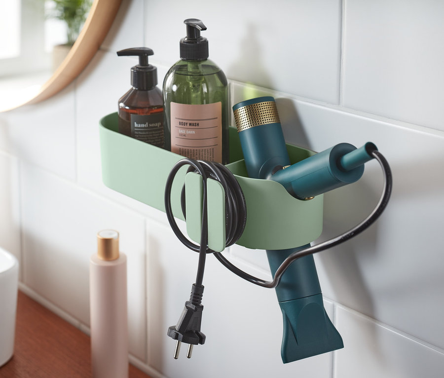 V koupelně je na bílých dlaždicích upevněn zelený koupelnový organizér s hnědým mýdlem, zelenou lahví a fénem.