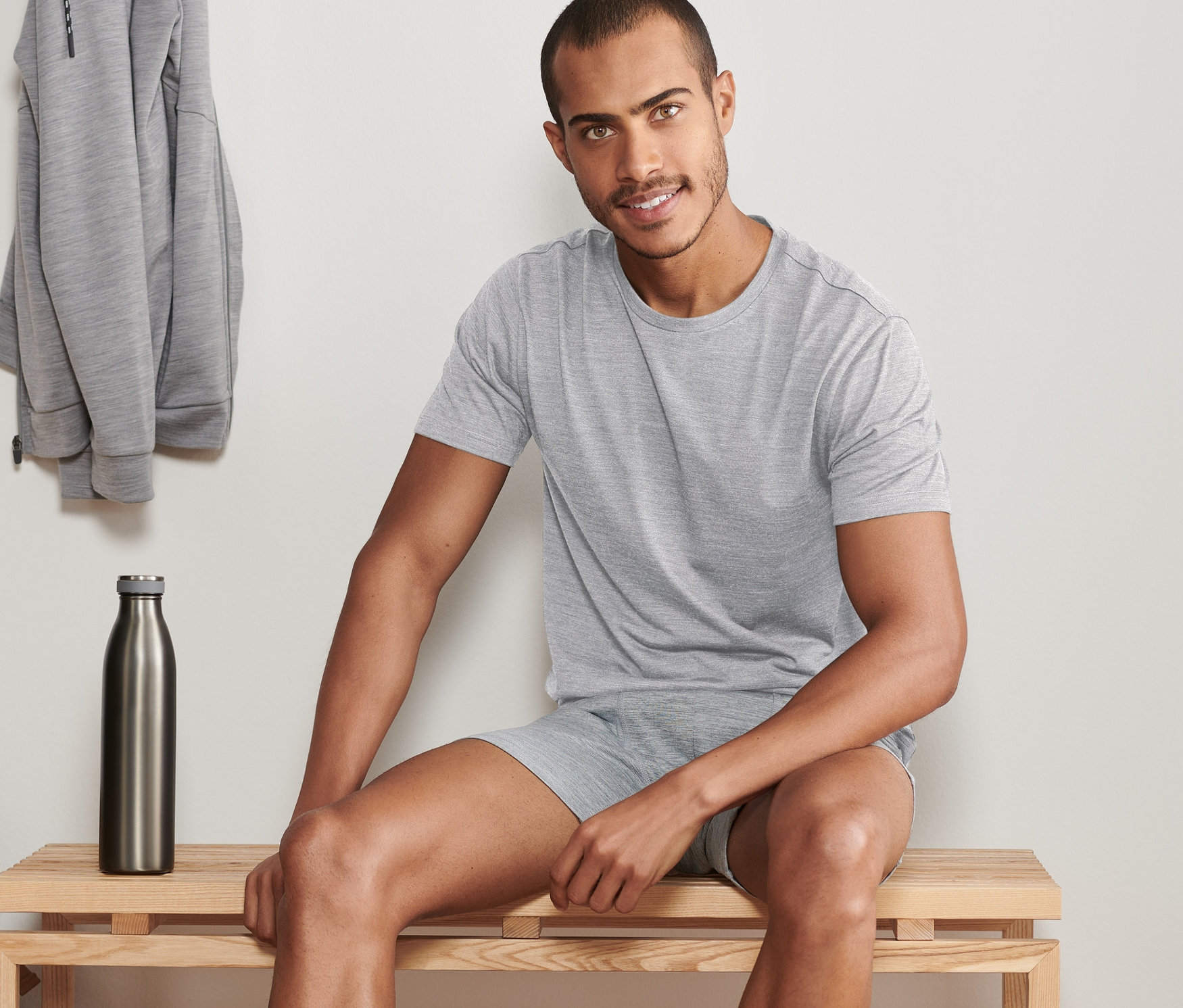 Muž sedí na lavici a má na sobě šedé funkční tričko. Na věšáku visí šedá mikina a na polici leží modrá helma. V pozadí je vidět láhev na vodu.