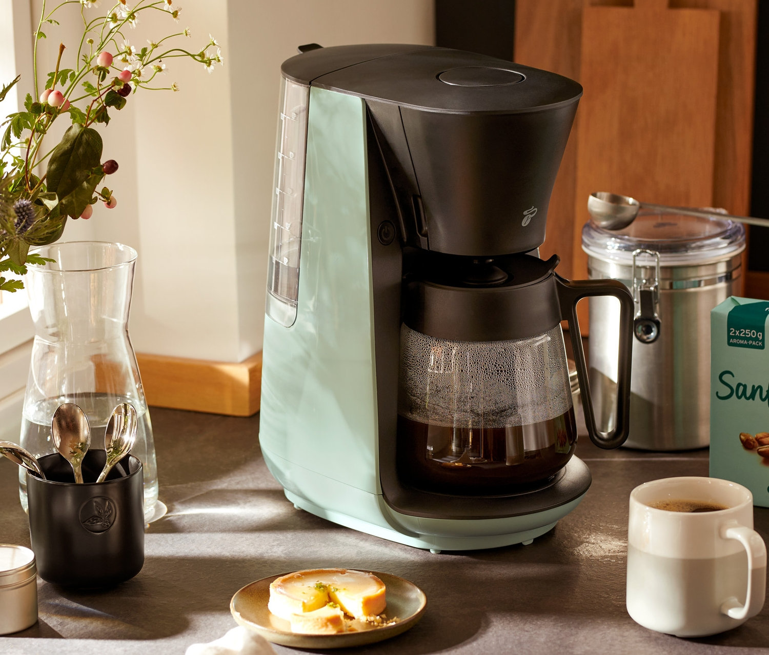 Světe zelený překapávač na kávu »Let's Brew« stojí na lince vedle šálku kávy a vázy s květinami.