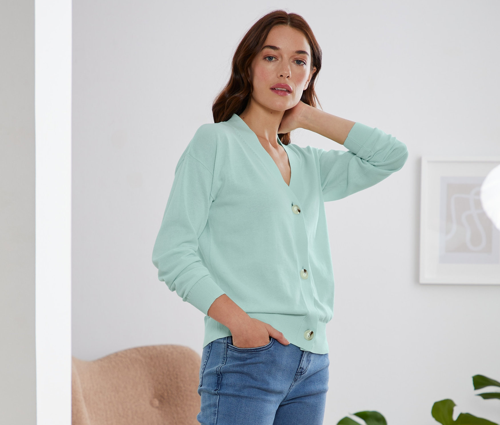 Žena pózuje v mátovém kardiganu s výstřihem do V, džínách a bílých teniskách.