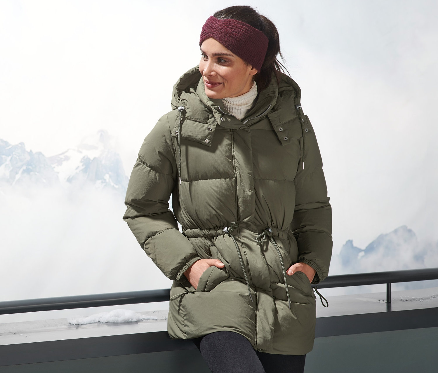 Žena v péřové bundě stojí venku s horami v pozadí.
