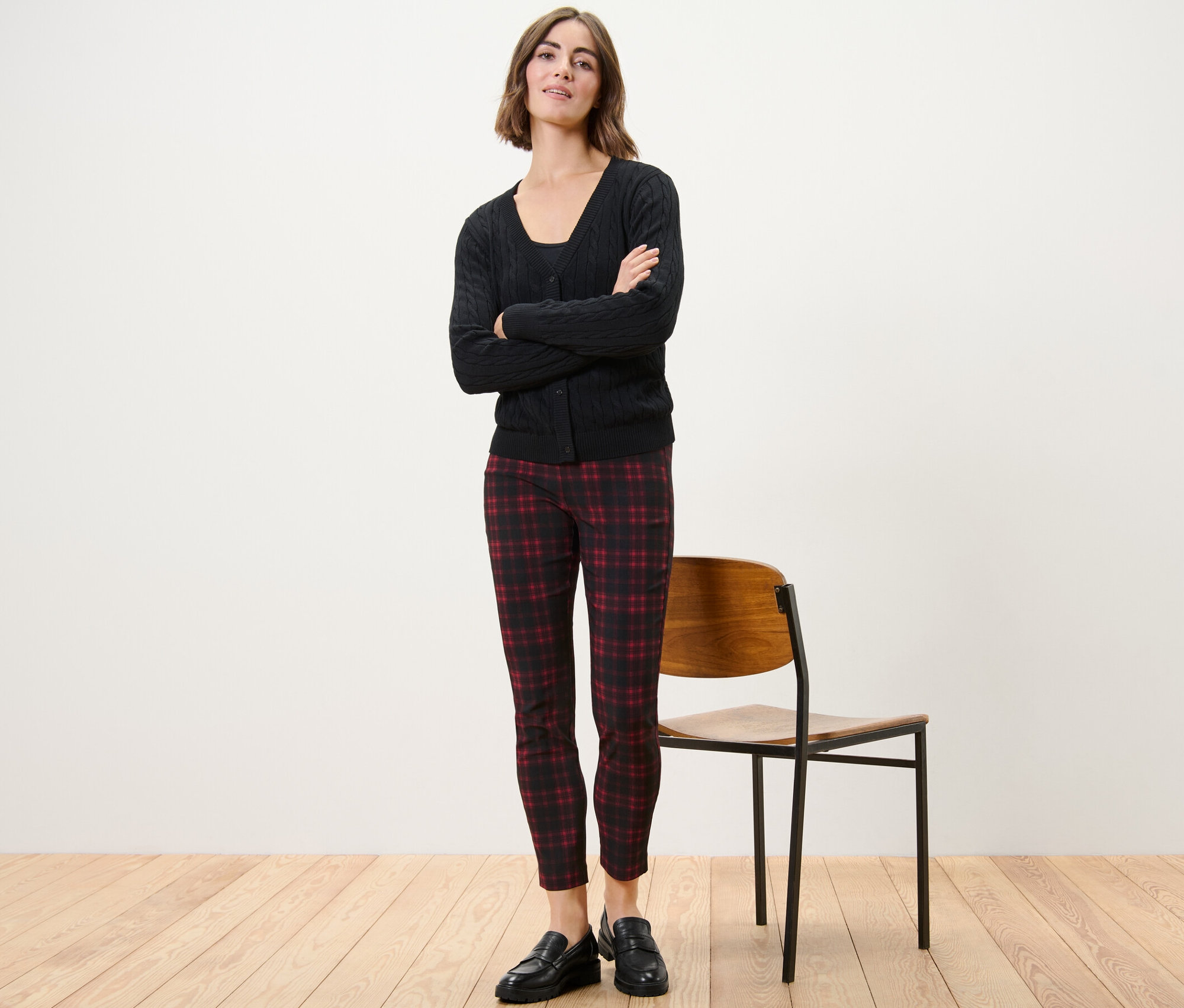 Žena stojí s rukama zkříženýma na prsou a má na sobě kostkované strečové kalhoty, černé sako a černé boty.