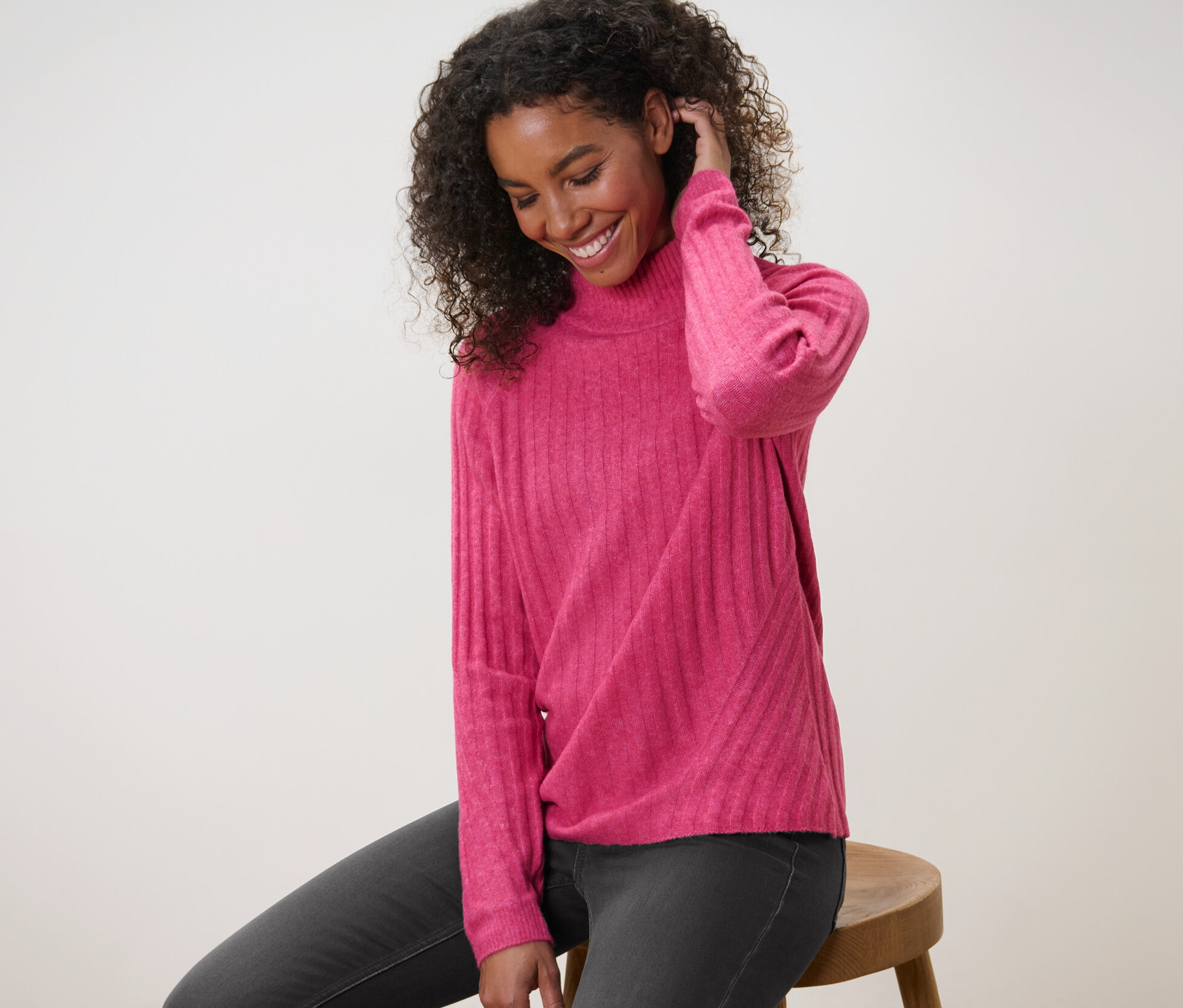 Žena sedí na stoličce a má na sobě růžový žebrovaný pletený svetr se stojáčkem.