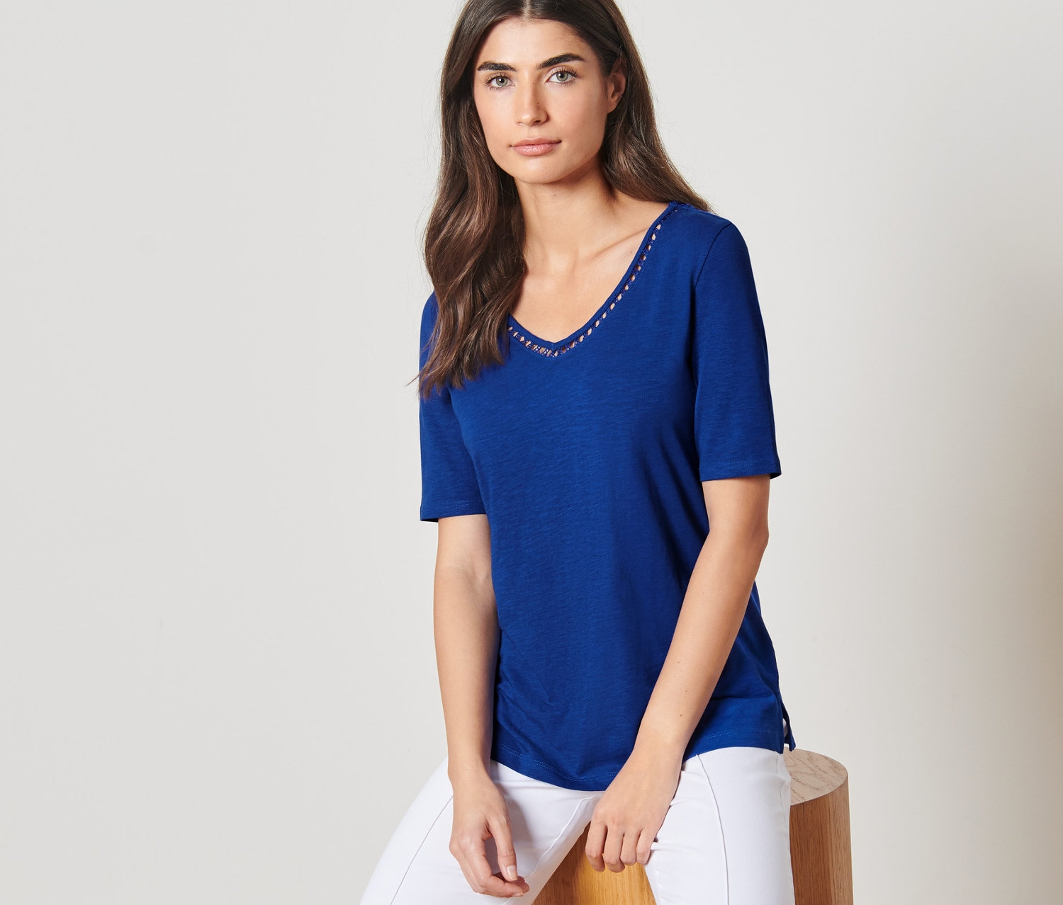 Žena sedí na stoličce a má na sobě kobaltově modrou košili a bílé kalhoty.