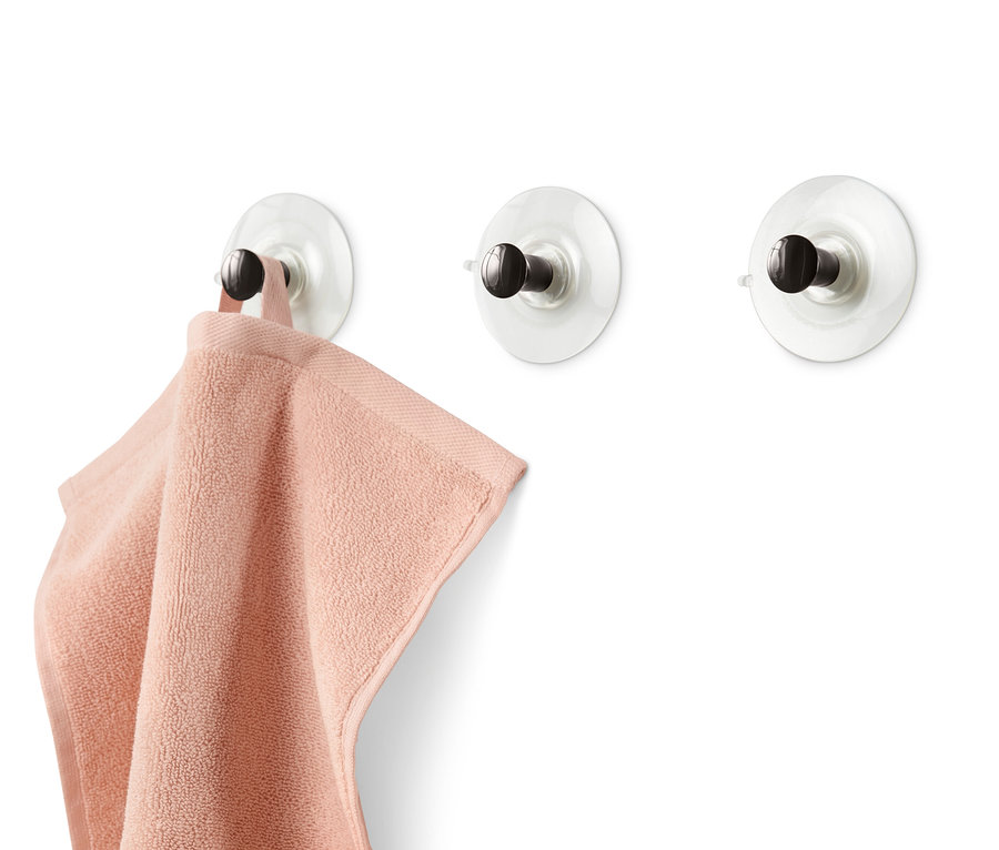 Na bílé zdi jsou připevněny 3 háčky s přísavkou, na jednom z nich visí růžový ručník.