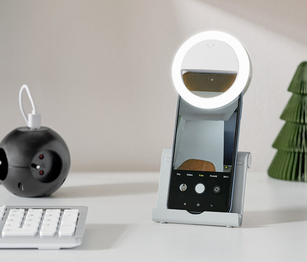 Stříbrný stojan na smartphone a tablet drží smartphone s kruhovým světlem na bílém stole.