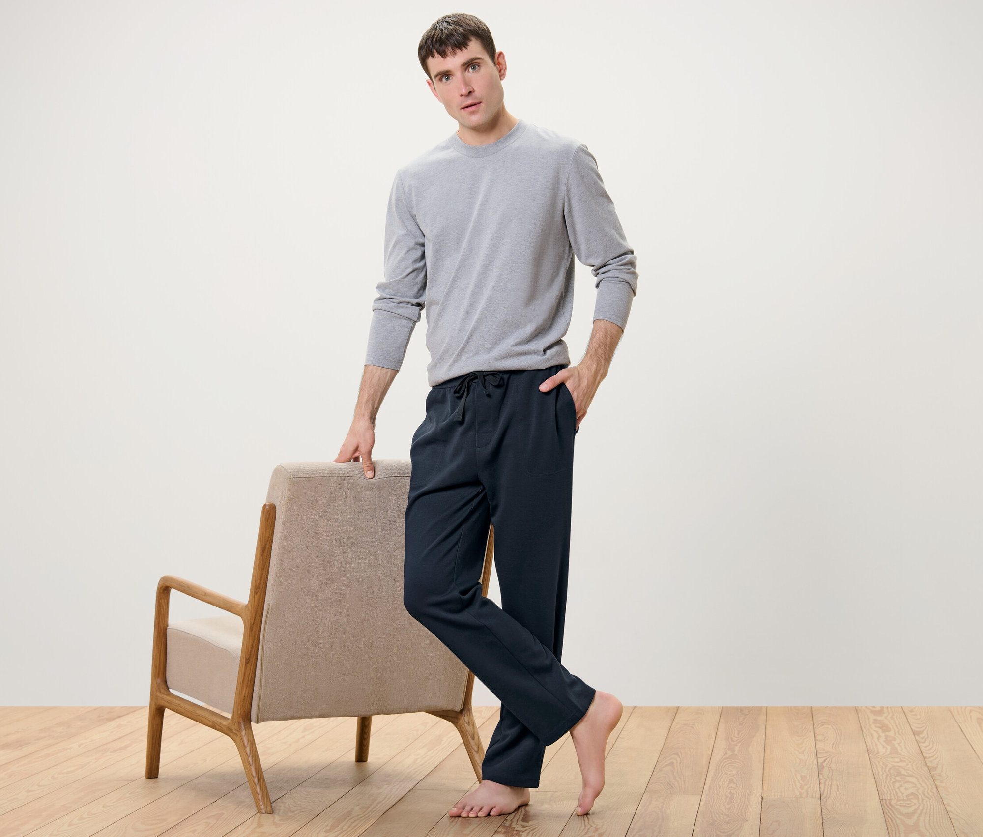 Muž pózuje vedle křesla a má na sobě domácí kalhoty a šedé tričko.