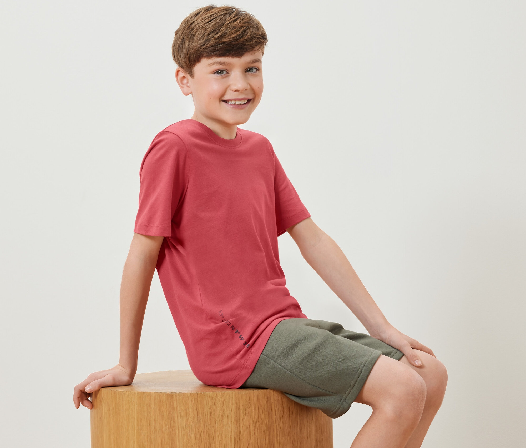 Chlapec sedí na dřevěné kostce a má na sobě jedno červené tričko ze sady Dětská trička, 2 ks.