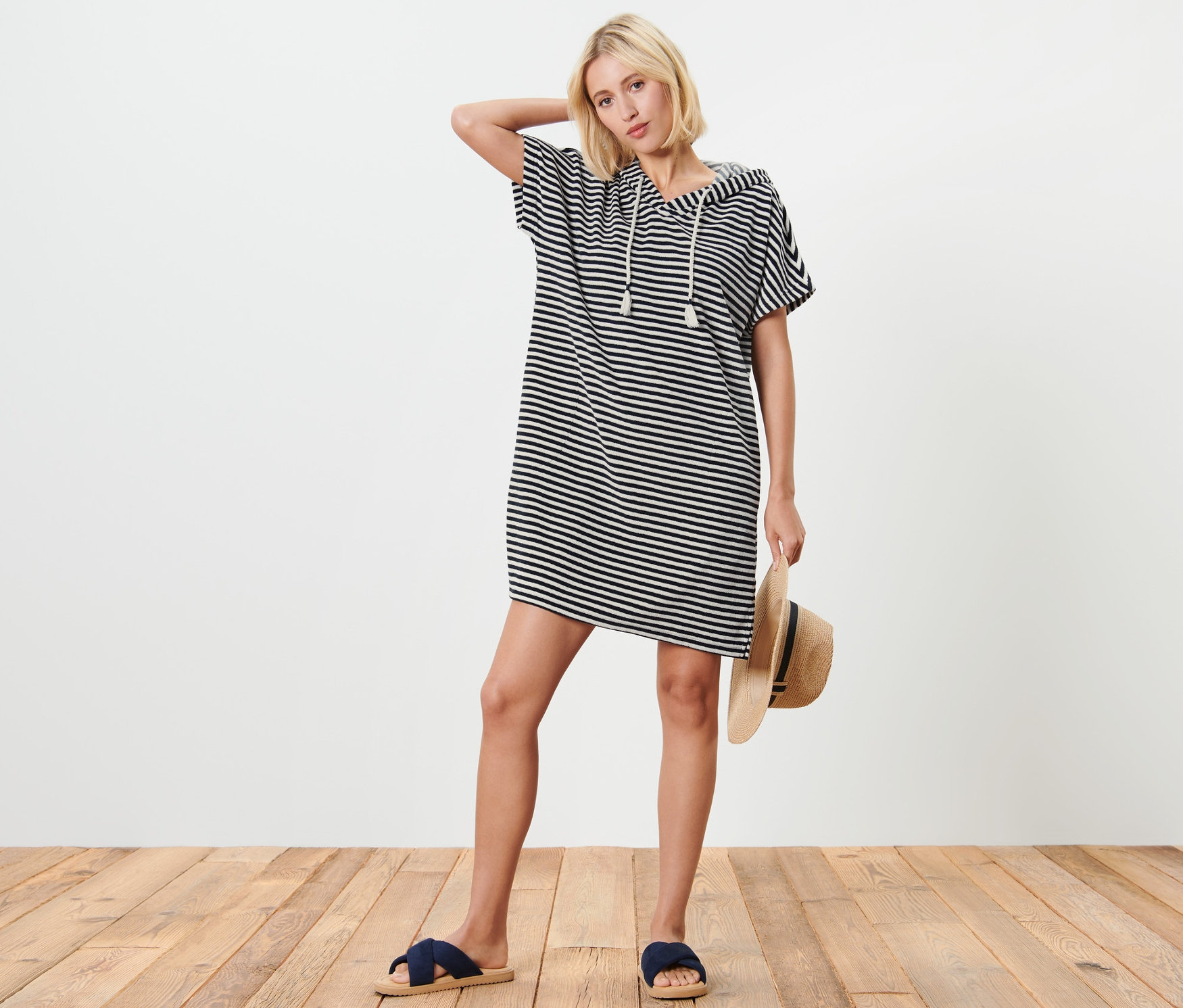 Blonďatá žena pózuje v modro-bílých plážových froté šatech s kapucí, drží slaměný klobouk a má na sobě modré pantofle.