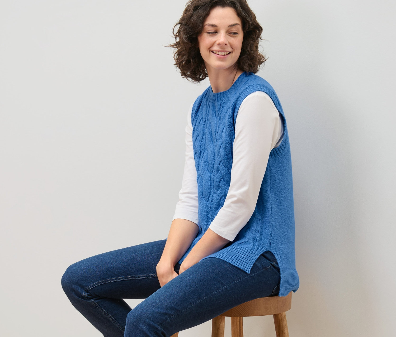 Žena sedí na stoličce a má na sobě pletenou vestu s copánkovým vzorem.