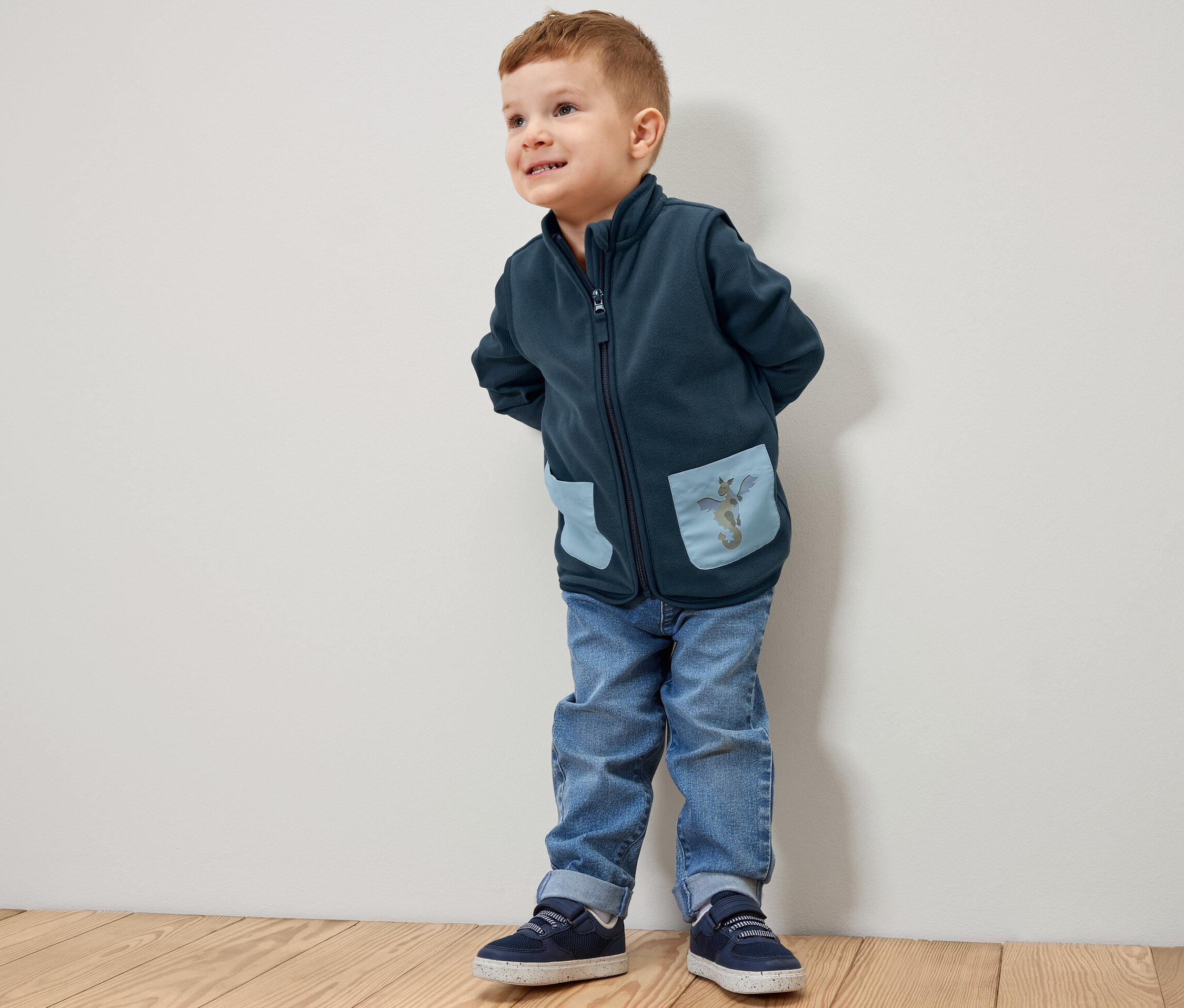 Chlapec stojí a má na sobě dětskou fleecovou vestu s motivem dráčka.