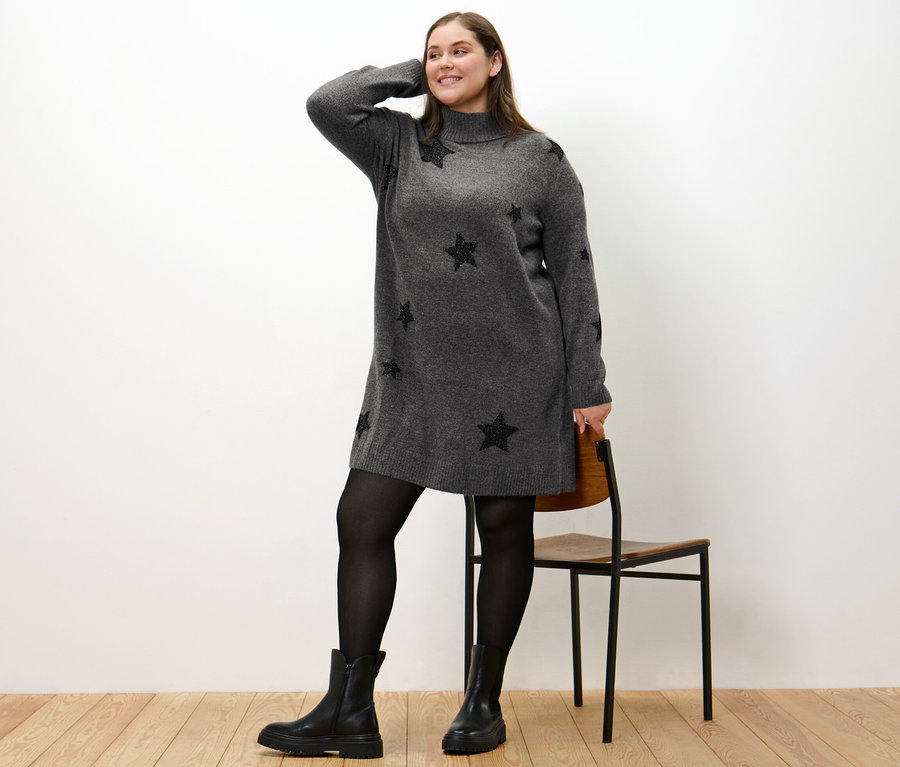Žena pózuje v pletených šatech s rolákem a černými hvězdami, má černé boty a punčocháče.