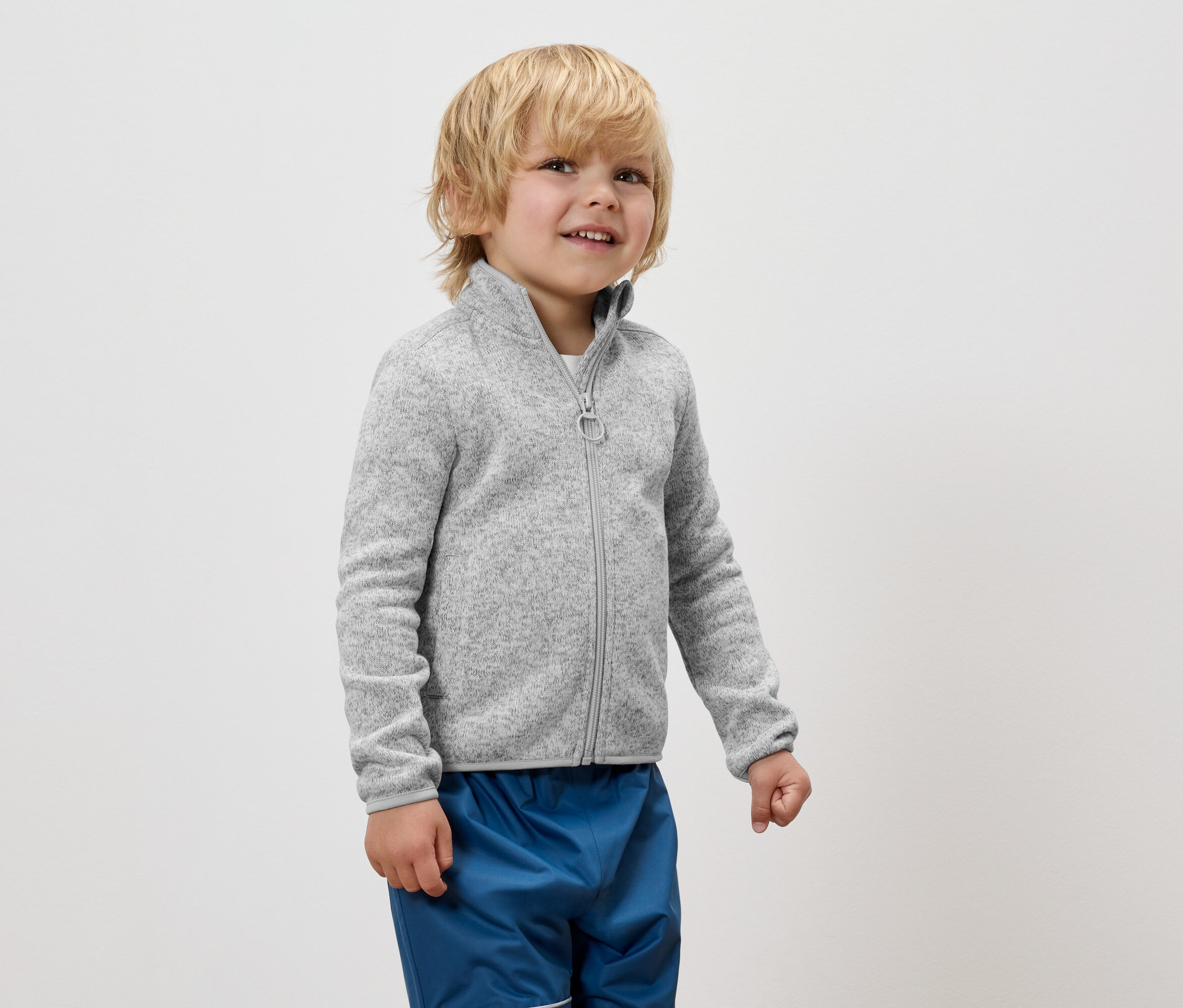 Chlapec v šedé mikině na zip a modrých kalhotách stojí na bílém pozadí.