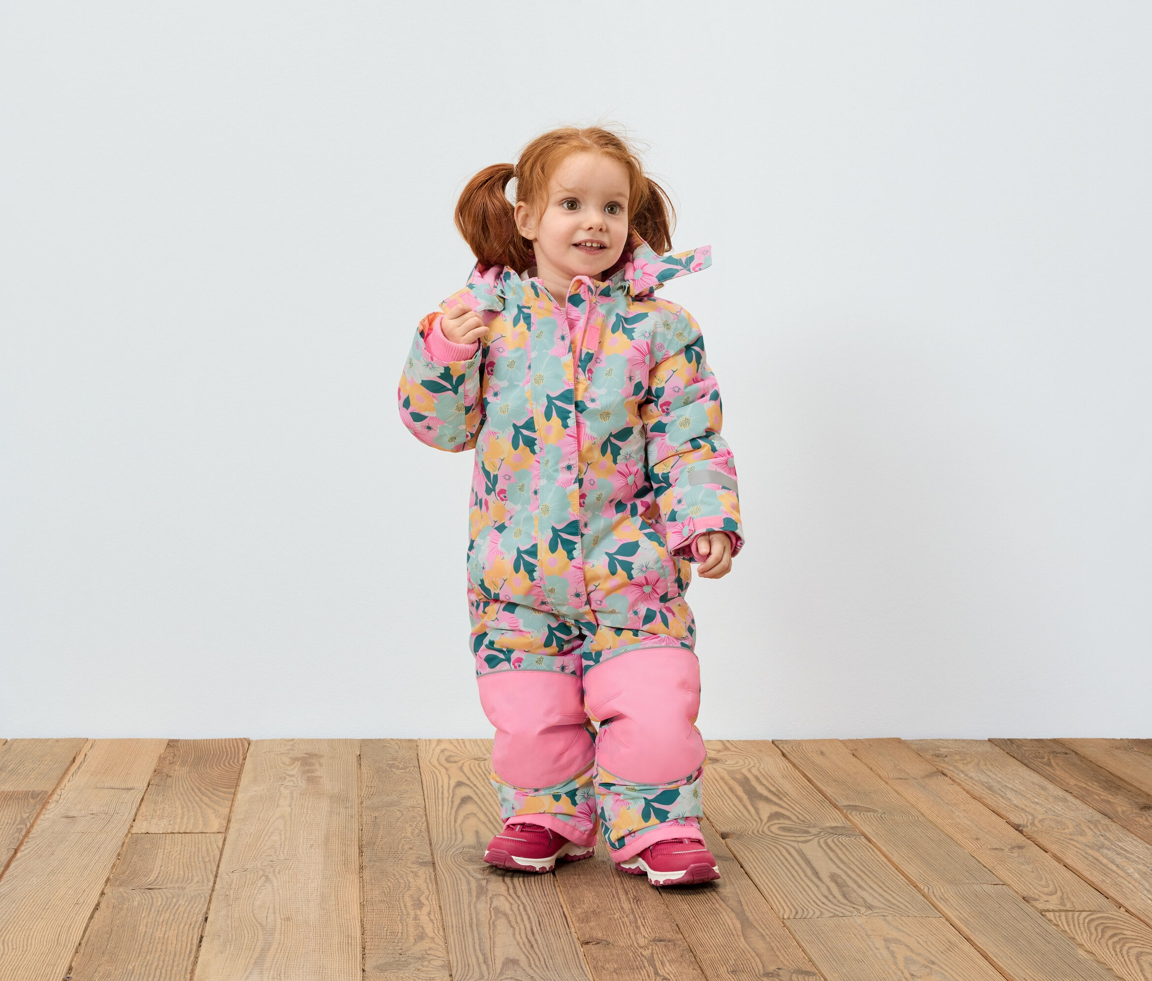 Dívka s rusými vlasy v květinové kombinéze a růžových botách stojí na dřevěné podlaze před bílou zdí.