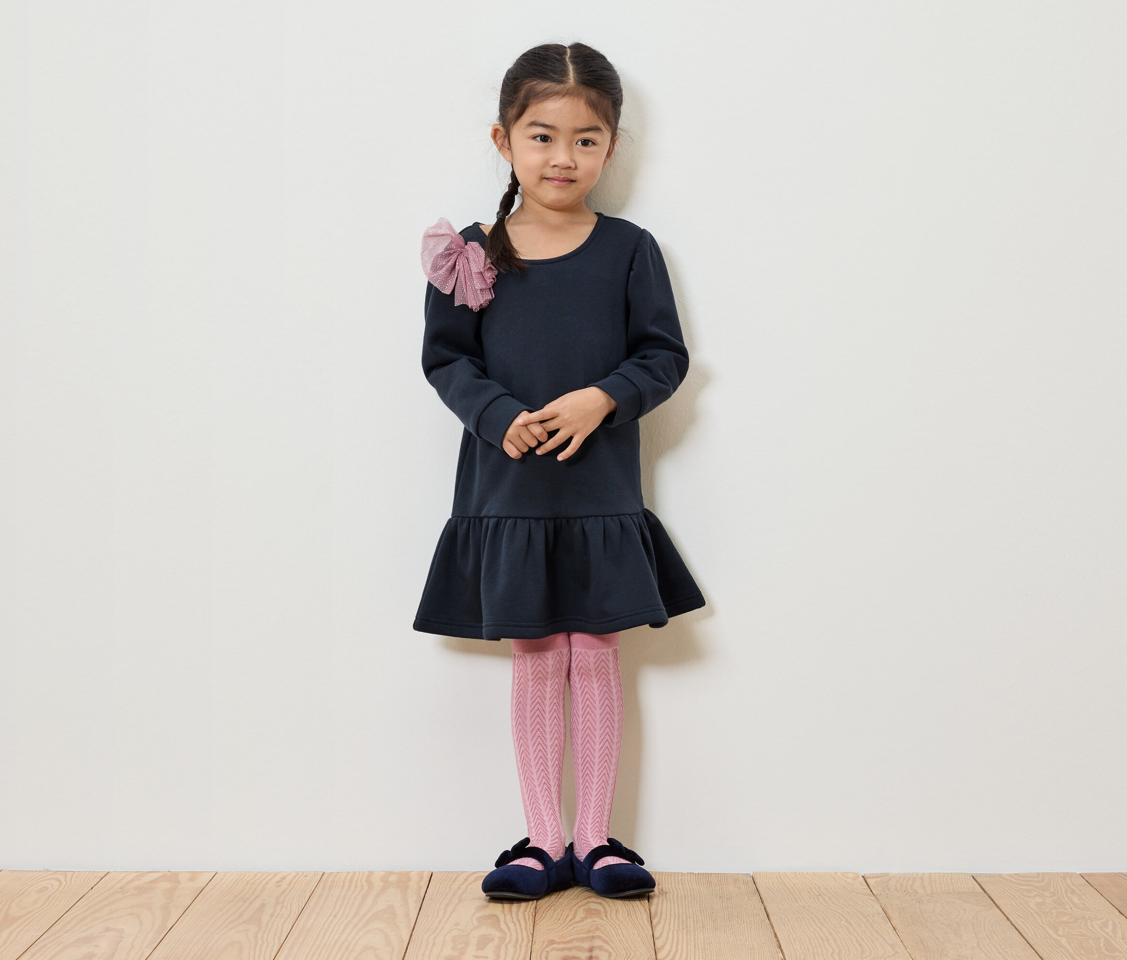 Dívka s copem stojí před bílou zdí. Má na sobě tmavě modré šaty, růžové punčocháče a modré boty.