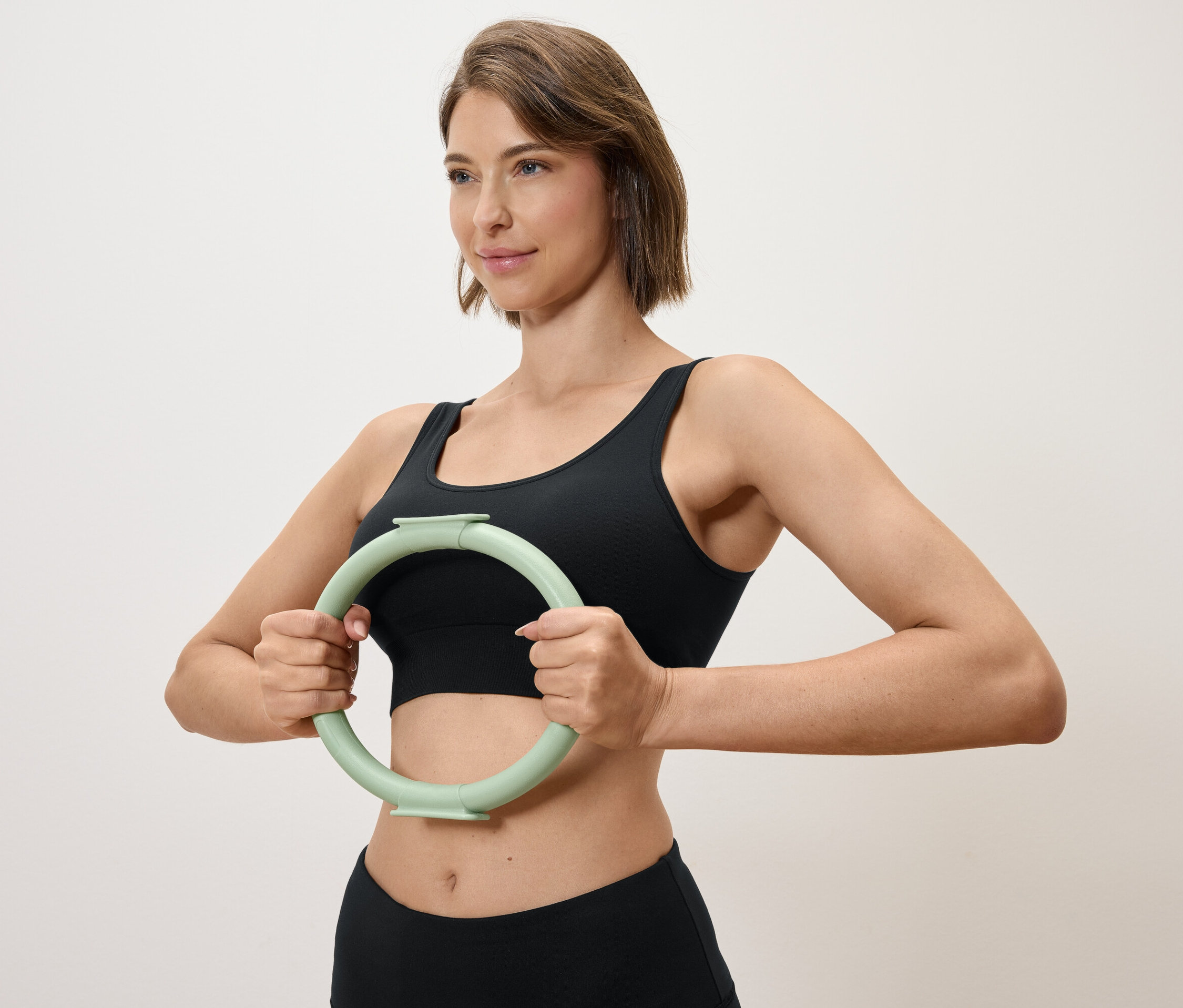 Žena drží světle zelený kruh na cvičení před sebou, oblečená do černého sportovního topu a legín.