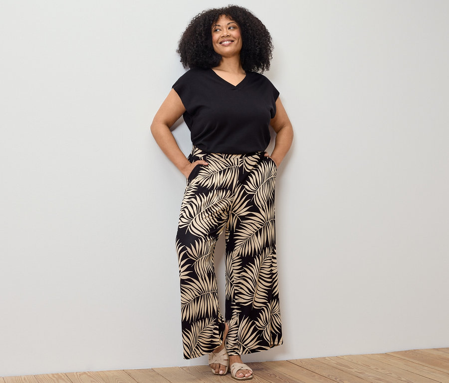 Žena pózuje v černém svetru z jemného úpletu s krátkým rukávem a vzorovaných širokých kalhotách.