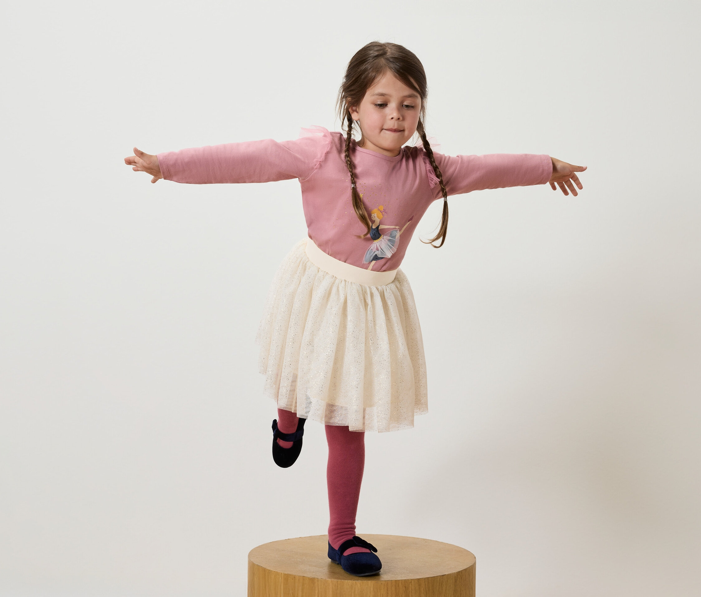 Dívka s hnědými vlasy v culících stojí na jedné noze na dřevěném podstavci, má růžové tričko, bílou sukni a tmavě modré boty.