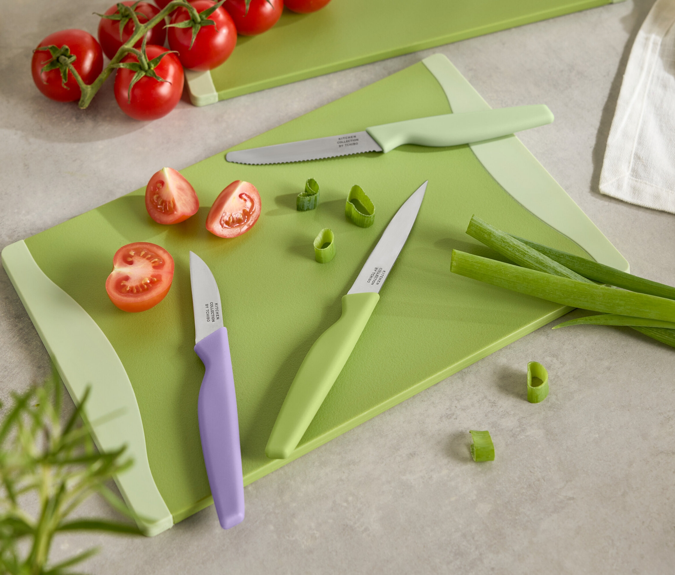 Zelené prkénko s nakrájenými rajčaty, jarní cibulkou a třemi noži s barevnými rukojeťmi.