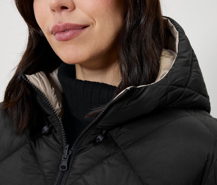 Detail dámy s černou prošívanou bundou s kapucí a černým rolákem.