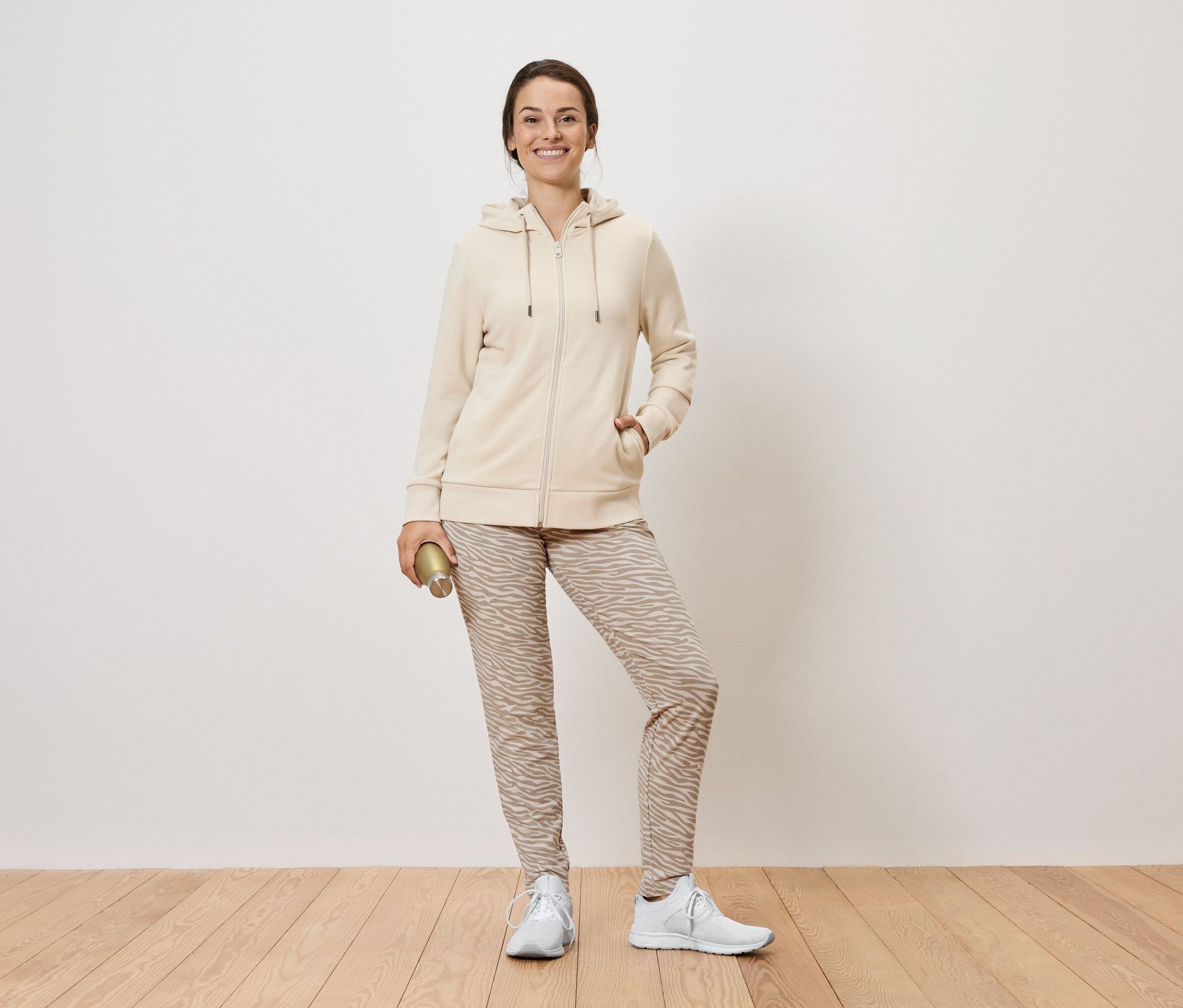 Žena stojí a drží zlatou láhev. Má na sobě béžovou mikinu na zip, vzorované kalhoty a bílé tenisky.