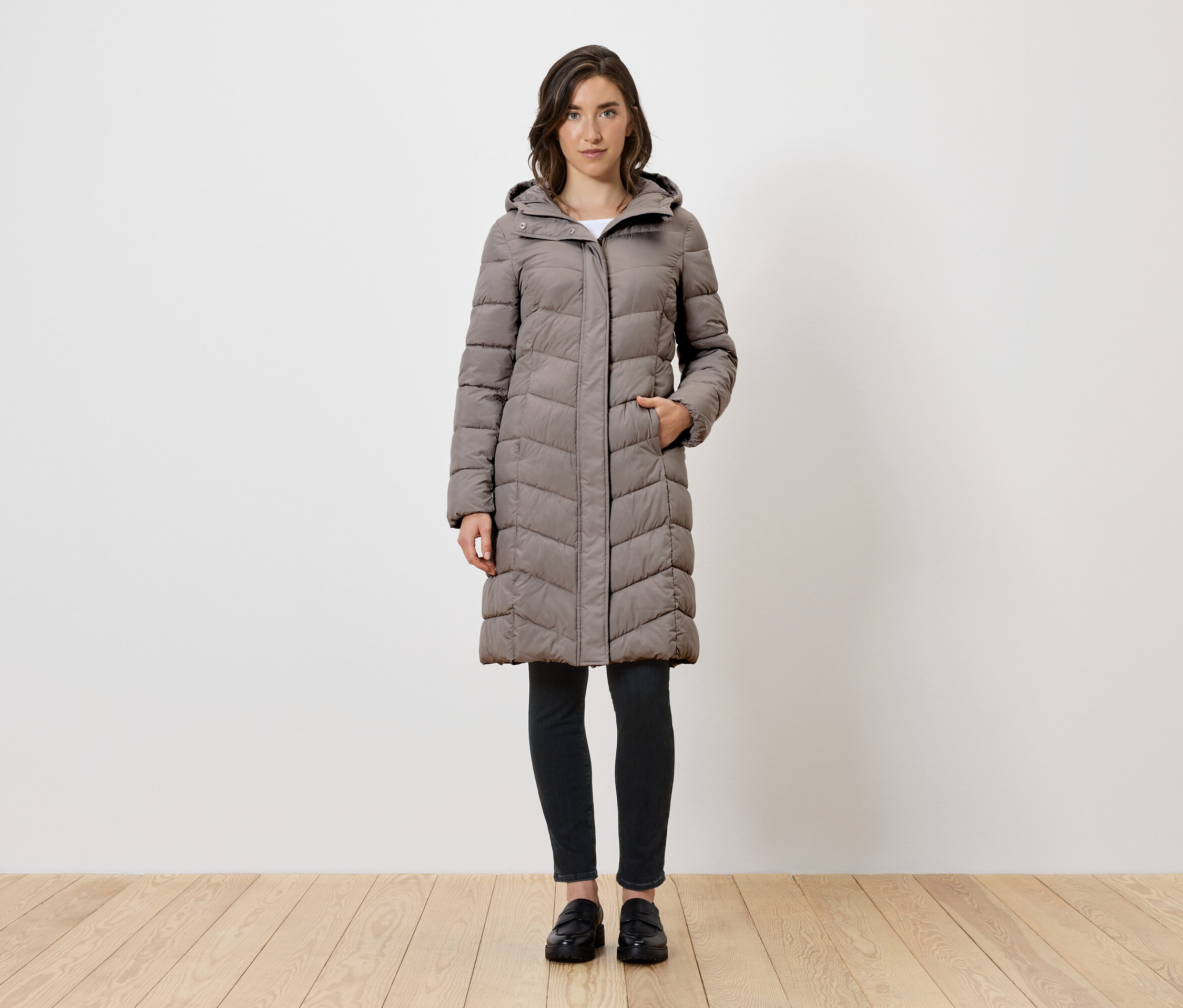 Žena stojí a má na sobě dlouhý šedý péřový kabát s kapucí, černé legíny a černé boty.
