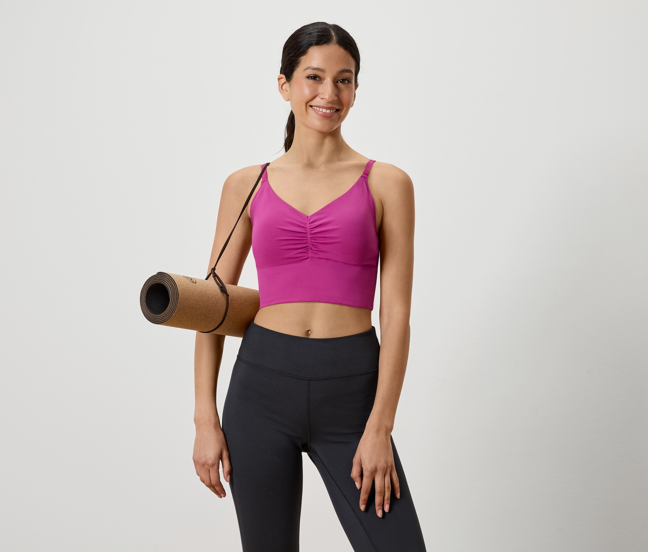 Žena s hnědou podložkou na jógu přes rameno, oblečená v růžovém sportovním topu a černých legínách.