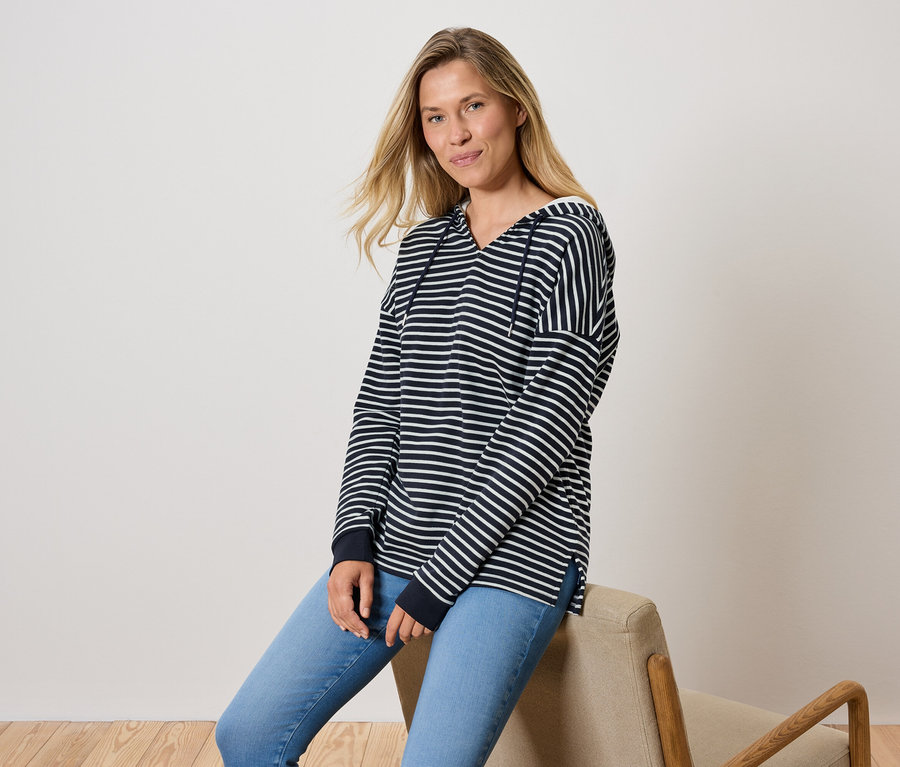 Žena sedí na židli, oblečená do modrých džínů a pruhované mikiny s kapucí.