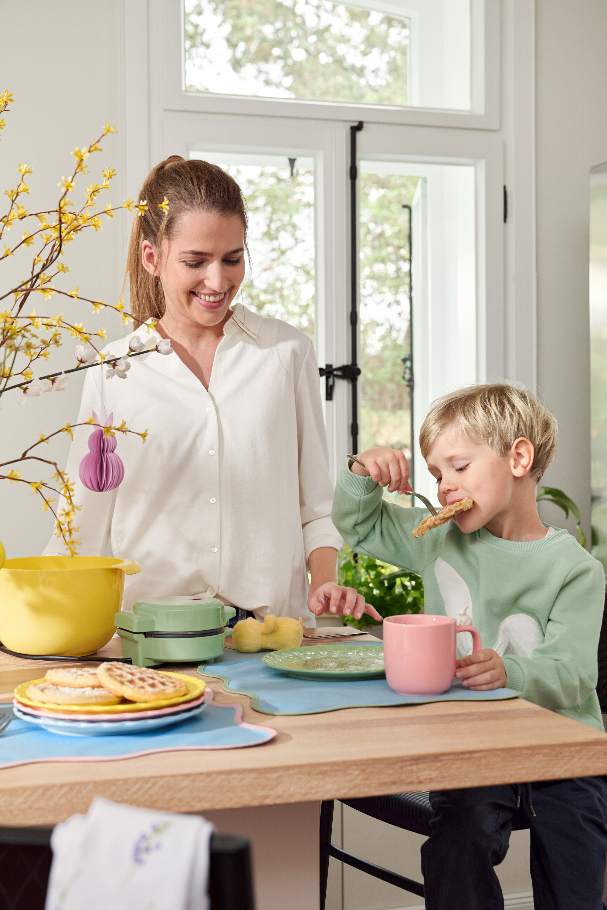 Žena a dítě u stolu. Dítě jí vafle vidličkou, žena se usmívá.