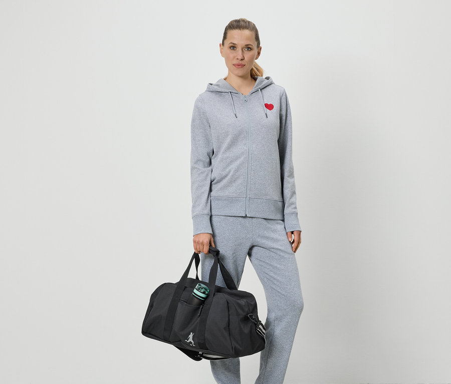 Žena v šedé mikině na zip s červeným srdcem a teplákách drží černou sportovní tašku.