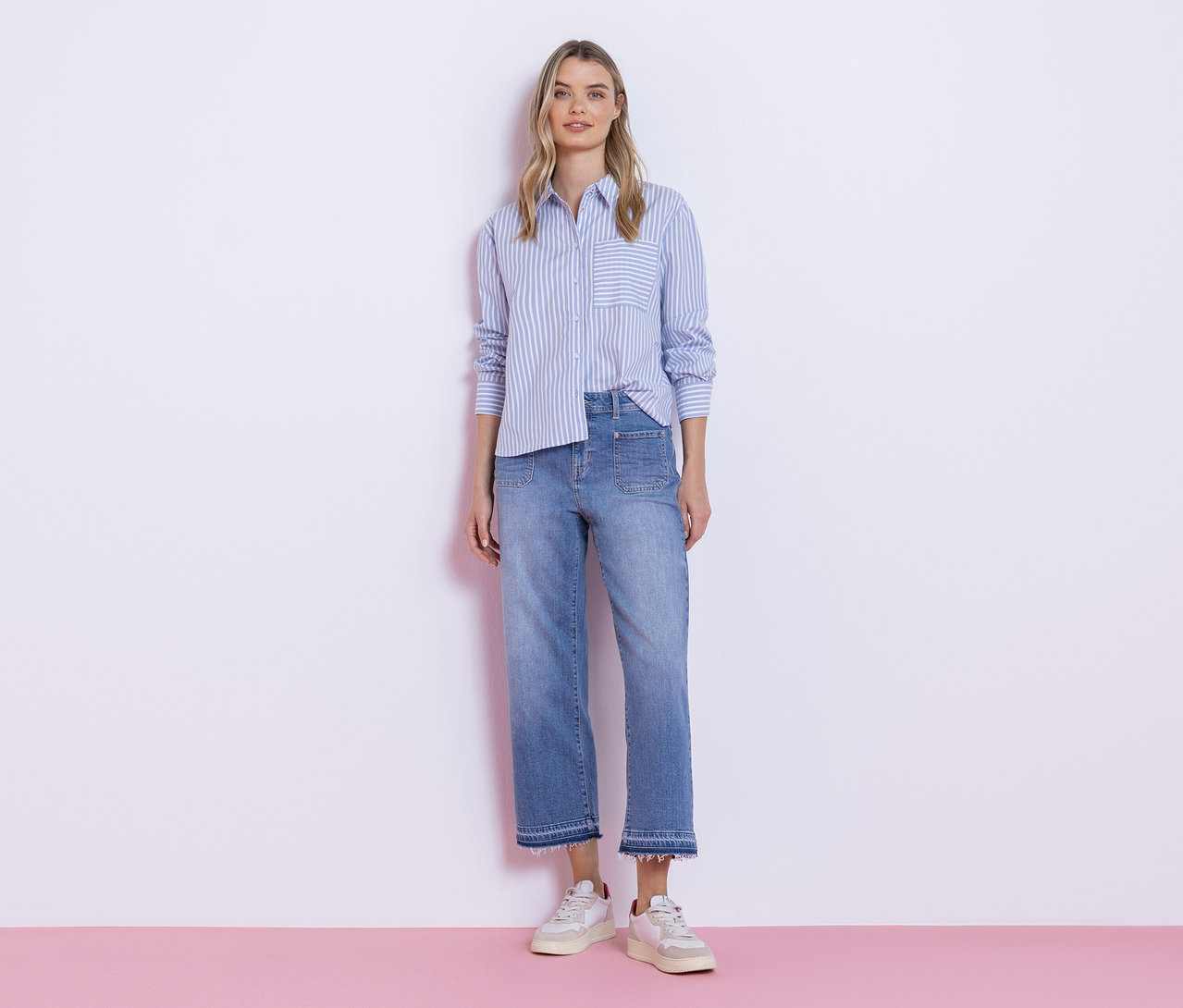 Mladá žena stojí před bílou stěnou, oblečená v modro-bílé pruhované košilové halence Street One, světlých džínách s roztřepeným lemem a bílých teniskách.