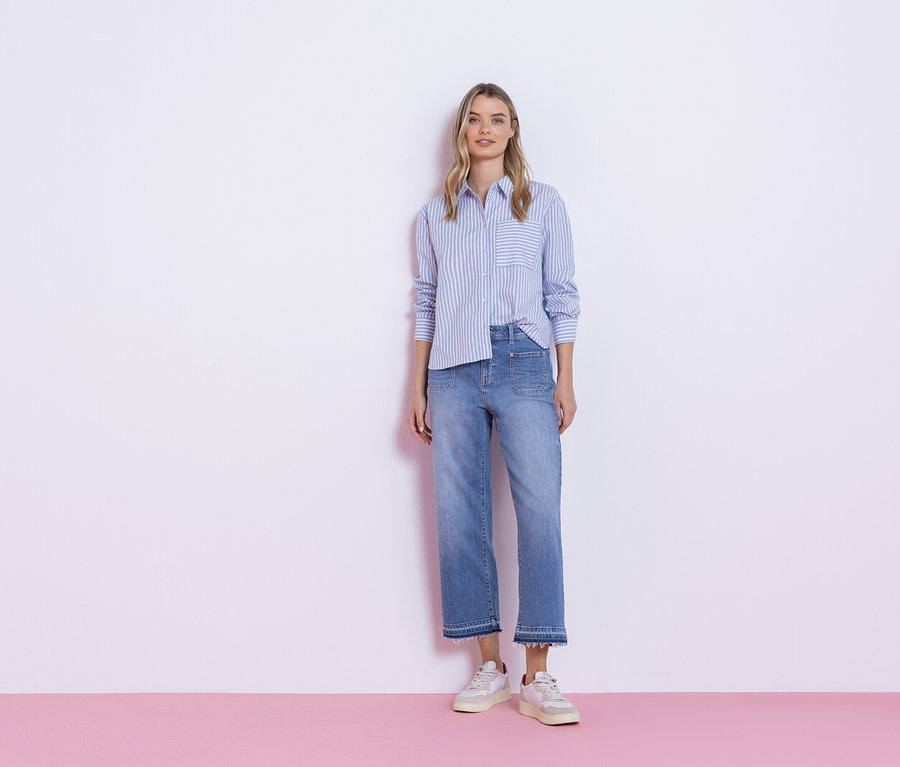 Žena stojí před bílou zdí, oblečená v modré pruhované košili, džíny a tenisky.
