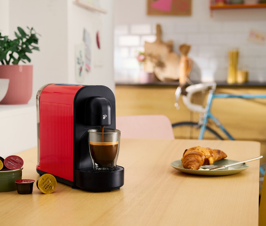 Červený kapslový kávovar plní šálek kávou. Na stole leží croissant, miska s kávovými kapslemi a hrnková rostlina.