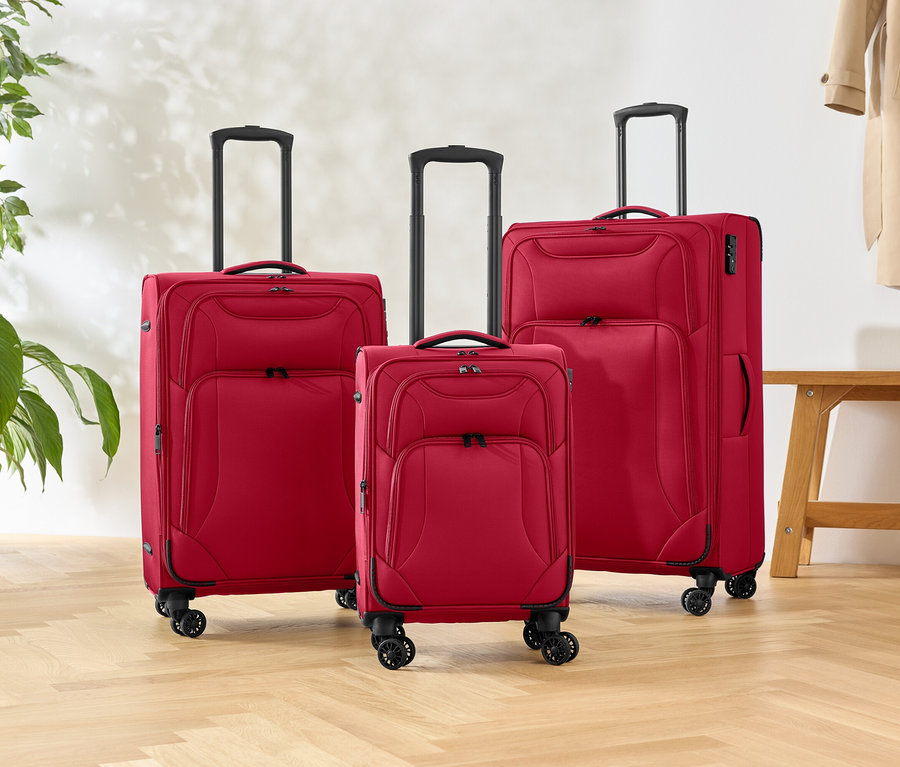 Tři červené kufry různých velikostí stojí na dřevěné podlaze, vedle rostliny a lavice.