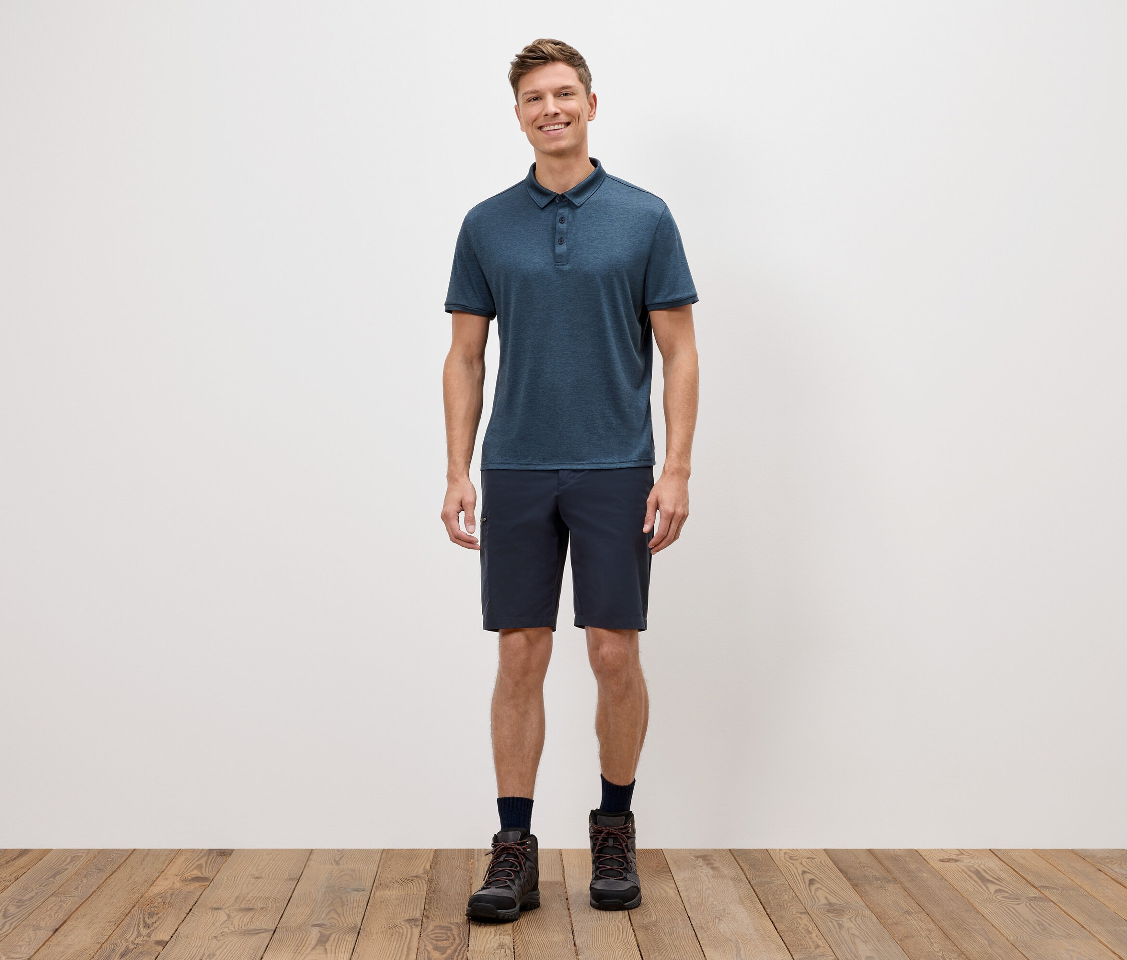 Muž stojí před bílou zdí a má na sobě modré polotriko, krátké šortky a trekové boty.