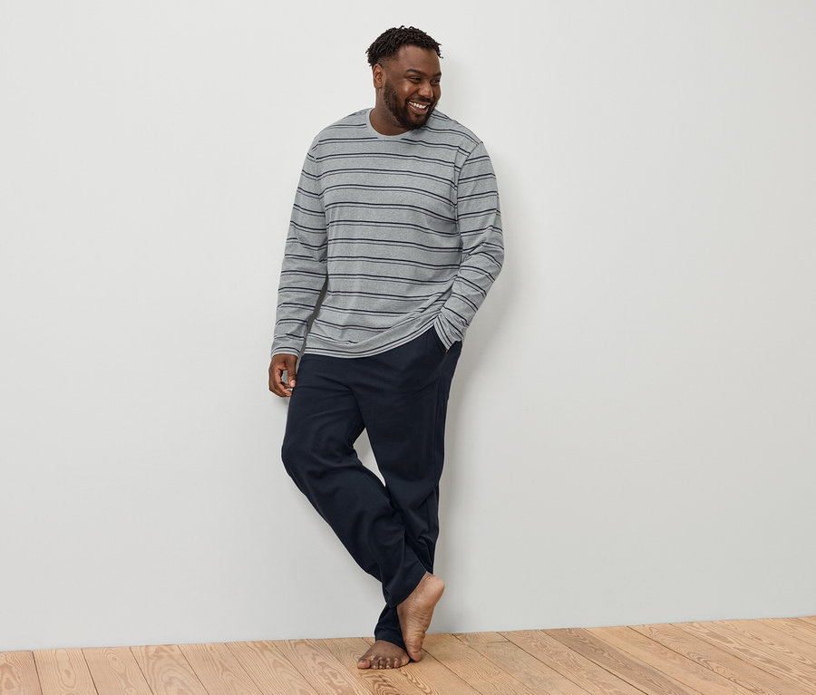 Usmívající se muž s tmavými vlasy a vousy stojí bosý, oblečený v šedém pruhovaném tričku s dlouhým rukávem a tmavě modrých kalhotách.