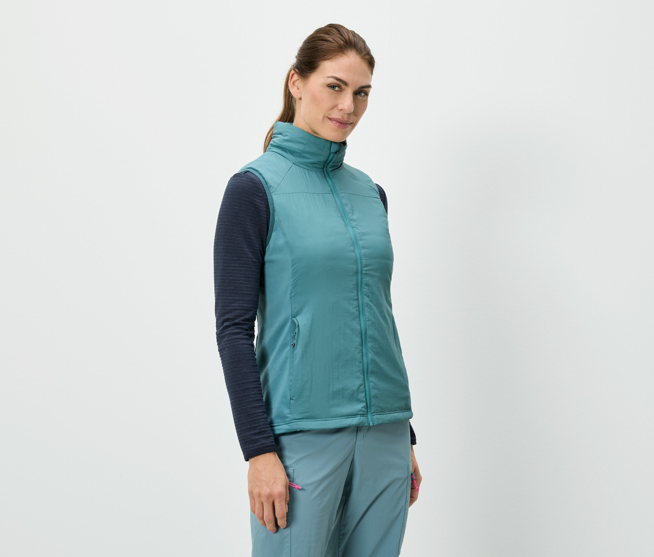 Žena stojí a má na sobě modrou vestu přes tričko s dlouhými rukávy, v kombinaci s modrými kalhotami.