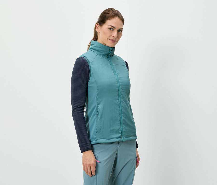 Žena stojí a má na sobě modrou vestu přes tričko s dlouhými rukávy, v kombinaci s modrými kalhotami.