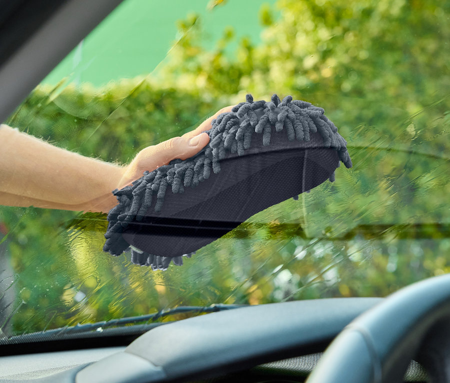 Ruka stírá čelní sklo auta šedou mycí houbou.