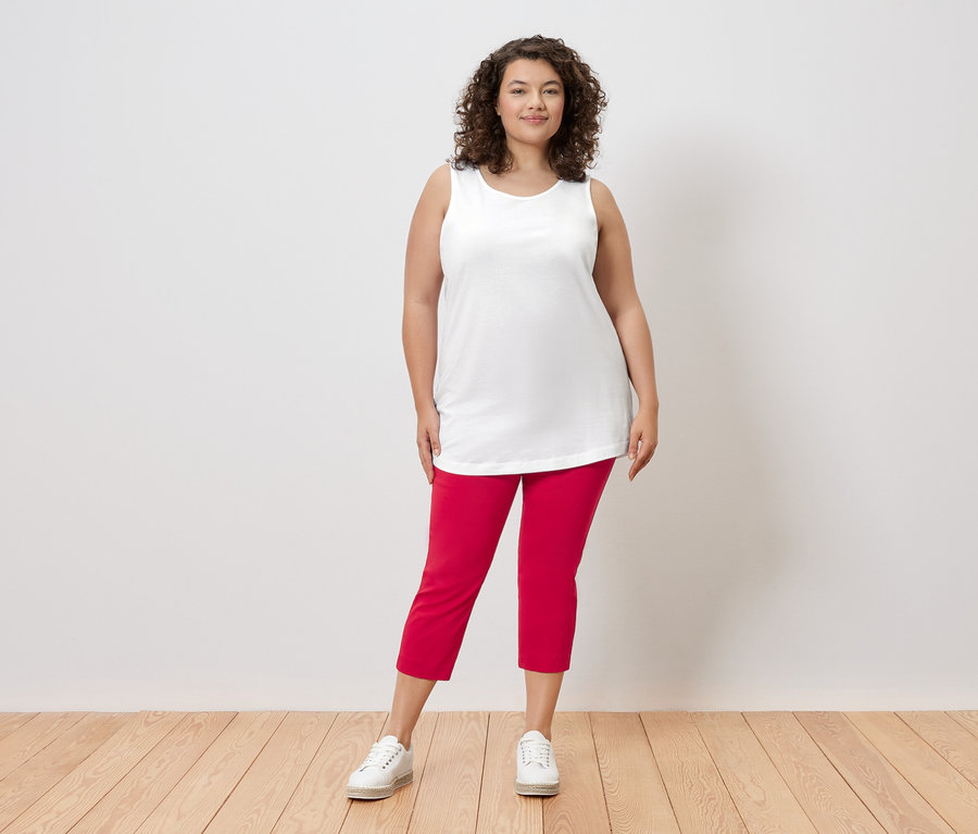 Žena s kudrnatými vlasy stojí před stěnou a má na sobě bílý top, červené kalhoty a bílé tenisky.