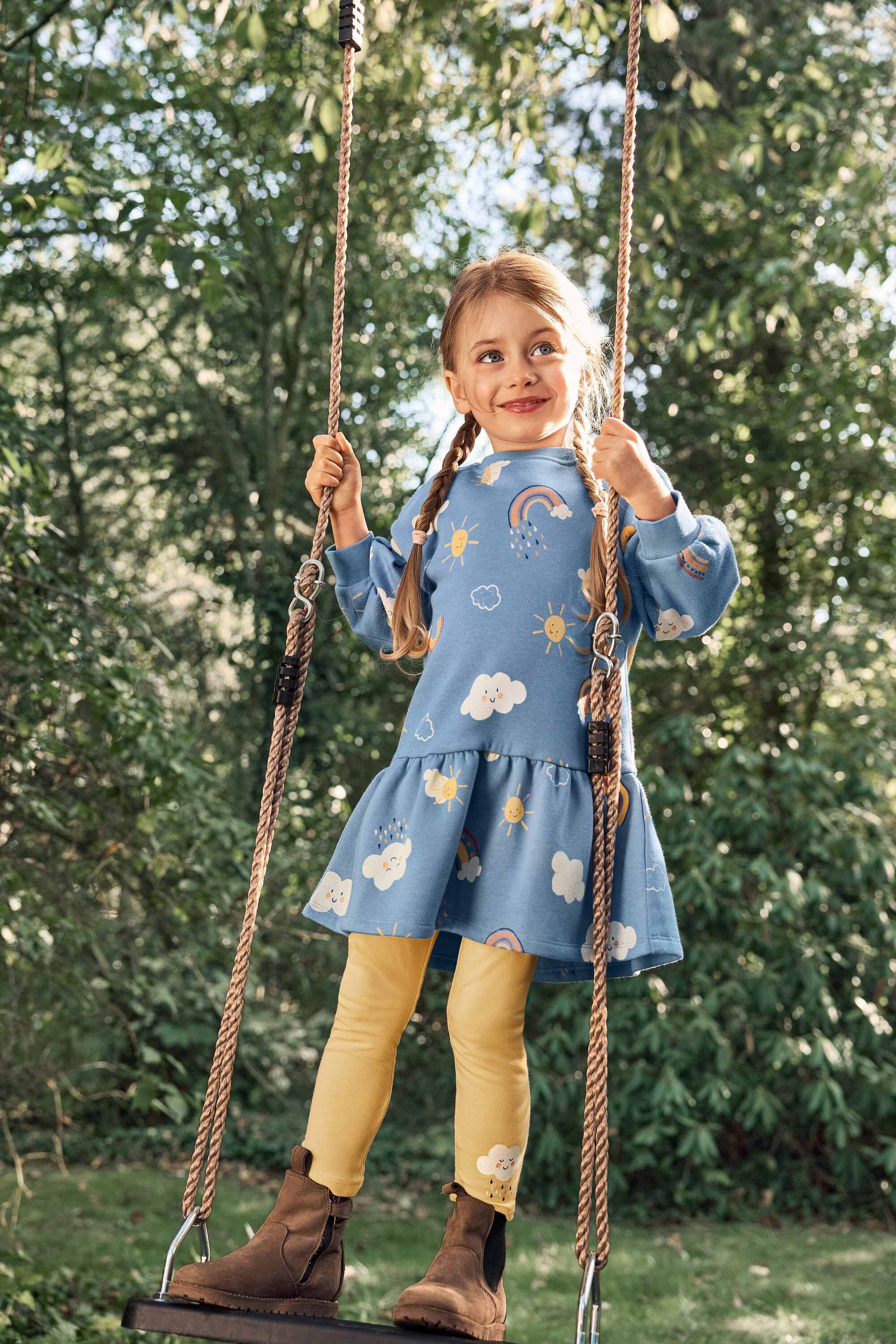 Dívka se zapletenými vlasy se houpe na houpačce, má na sobě modré šaty se vzorem slunce, mraků a duhy, žluté legíny a hnědé boty.