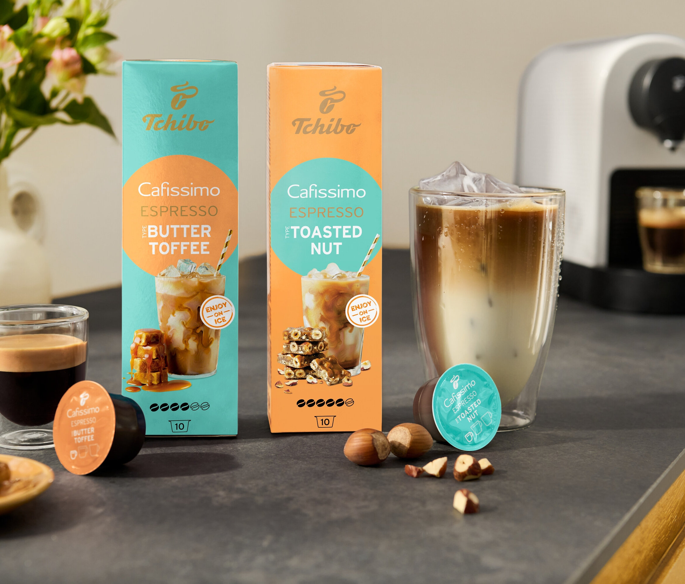 Na kuchyňské lince stojí dvě balení Cafissimo Flavoured Espresso, jedno v tyrkysové barvě s příchutí Buttertoffee a druhé v oranžové barvě s příchutí Toasted Nut, vedle bílého kávovaru Cafissimo pure+.