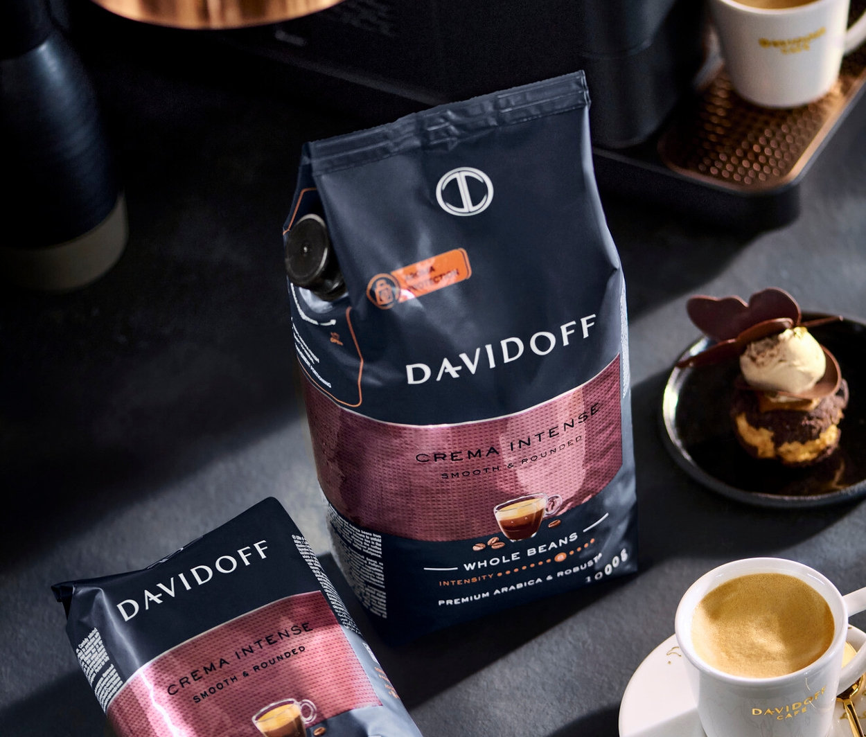 Dvě balení kávových zrn Davidoff Crema Intense, šálek kávy a dezert na stole.