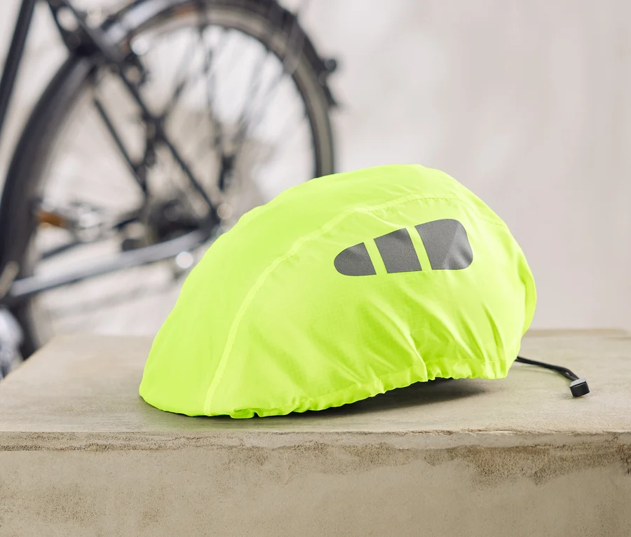 Neonově žlutý potah na cyklistickou helmu s reflexními pruhy leží na betonovém podstavci, v pozadí kolo.