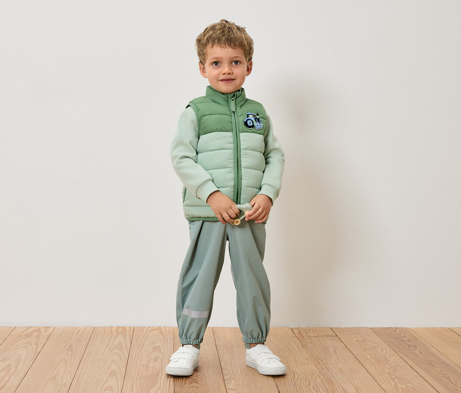 Chlapec stojí u zdi v světle zelené bundě, světle zelených kalhotách a bílých teniskách.